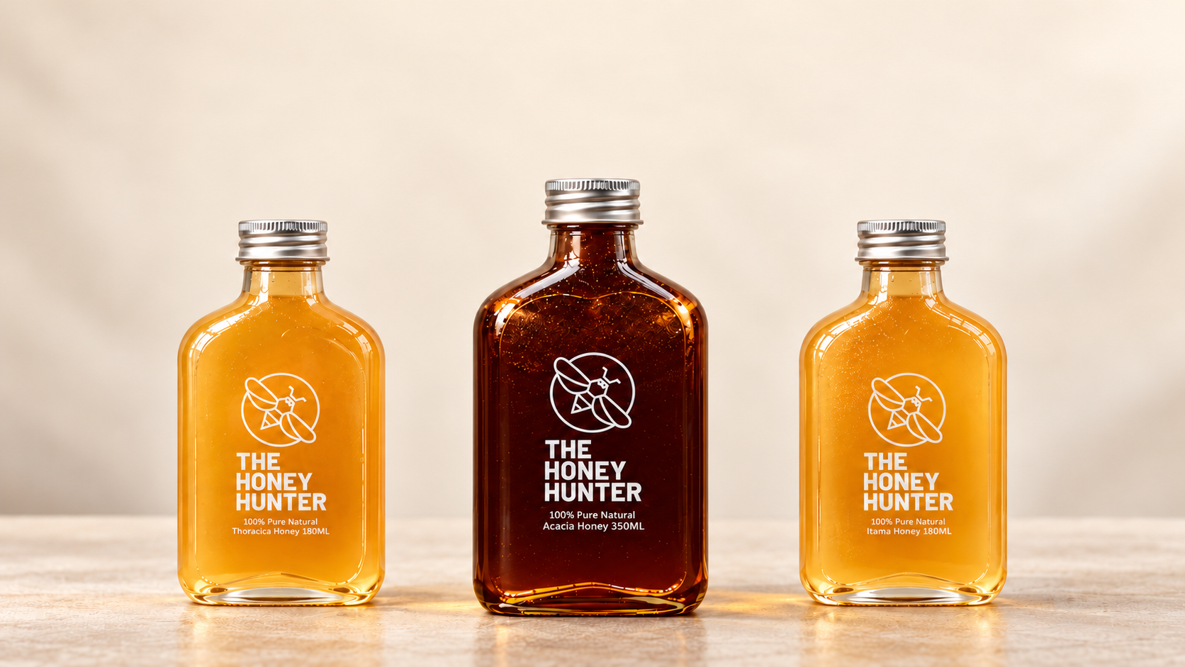 Three bottles of The Honey Hunter honey on a tabletop: Kelulut Thoracica on the left, the larger dark Acacia bottle in the centre and Kelulut Itama on the right.